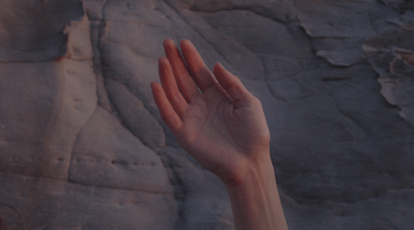 A hand with the palm facing forward and fingers slightly apart, set against a backdrop of a rock surface with natural lines and crevices. The lighting is soft and dim, suggesting either early morning or late afternoon, with warm tones that give the skin a reddish hue, contrasting with the cooler tones of the rock. The focus is on the hand, which is centered in the frame, while the rock surface in the background is slightly blurred. The composition is simple and evocative.