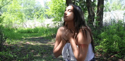 A person with long dark hair wearing a white sleeveless top and a patterned skirt is standing in a wooded area with lush greenery. They have their eyes closed and hands clasped over their chest in a gesture that suggests contemplation or serenity. Sunlight filters through the trees, highlighting the natural setting and the individual's peaceful pose.