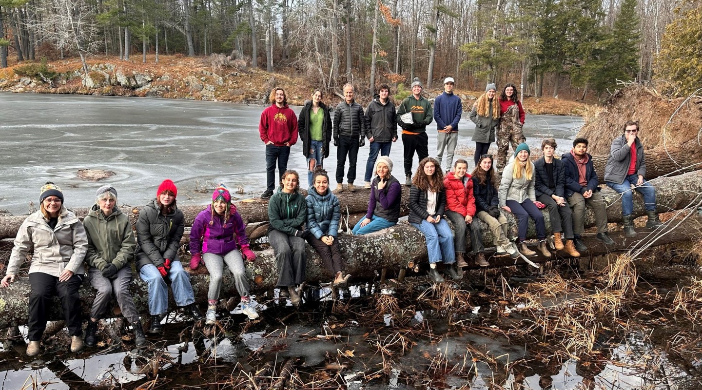 A group of twenty-two individuals of various attire are seated and standing on a fallen tree trunk over a body of frozen water, likely a lake or pond. The setting appears to be a cold day, as many individuals are wearing jackets, hats, and gloves. Bare trees and evergreens are visible in the background, suggesting a winter or late autumn season. The lighting suggests either early morning or late afternoon. There is no visible snow, but the ice on the water indicates freezing temperatures.
