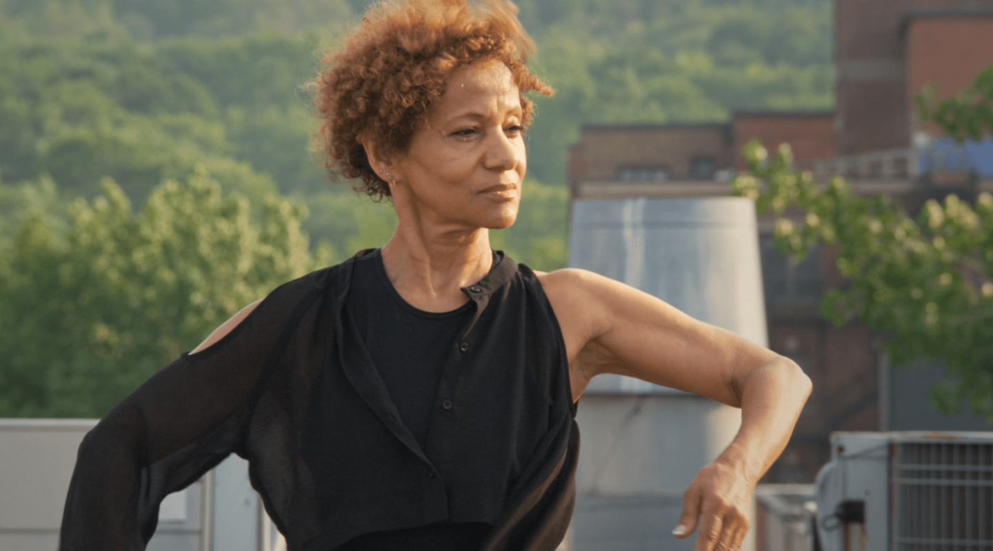 An individual with short curly hair is posing with one arm extended, wearing a black sleeveless top. They are outdoors, with a backdrop of greenery and urban structures, possibly on a rooftop. The person's expression is poised and serene, and the lighting suggests it could be late afternoon or early evening. The scene conveys a sense of calm confidence and the outdoors setting provides a relaxed atmosphere.