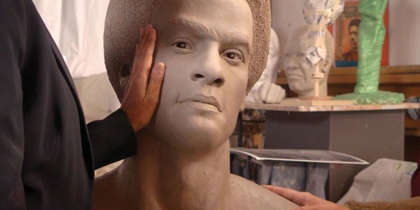 A person's hand is gently touching the cheek of a large, unfinished sculpted head and shoulders. The sculpture is highly detailed, with lifelike facial features, and it is mounted on a stand. In the background, there is an assortment of artistic materials and another bust sculpture, indicating that the setting is likely an artist's studio.