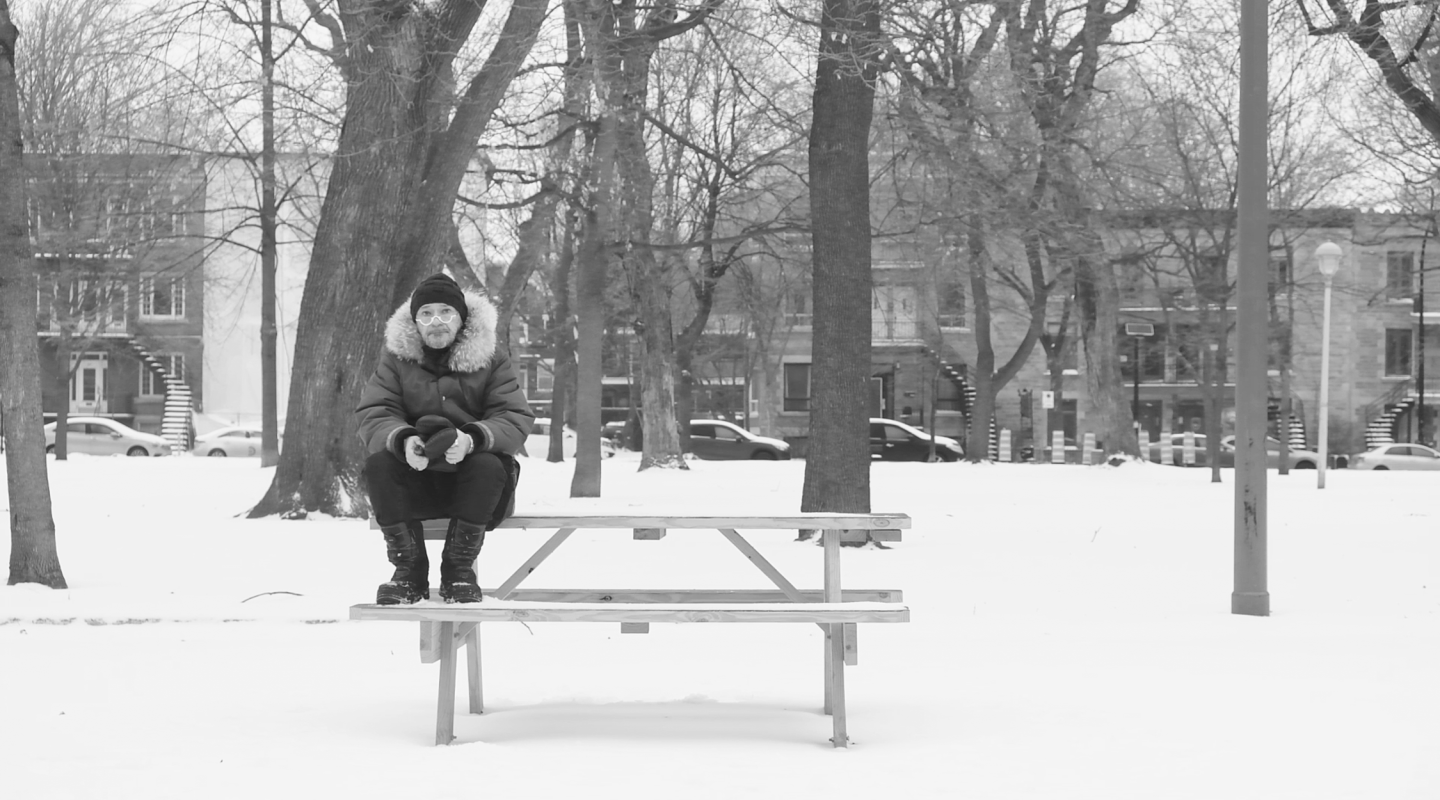 A black and white image of a snowy park setting, an individual is seated alone on a picnic table, dressed warmly in a heavy coat with a fur-lined hood, gloves, and a winter hat. The background features leafless trees, indicating it's a cold season, and buildings that suggest an urban environment. The person's posture and the empty park convey a sense of solitude or reflection.