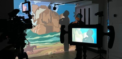 This image shows a behind-the-scenes view of a video production set. A video camera is in the foreground, focused on a person who is backlit against a large screen displaying colourful, stylized imagery resembling a landscape. The person's silhouette is also visible on the screen, indicating they might be performing or presenting. Monitors attached to the camera display the same image, allowing the crew to see what is being captured.