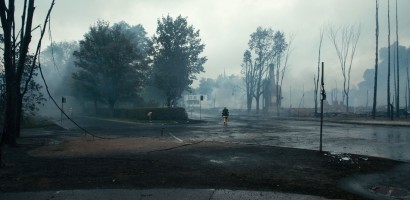A firefighter is walking through a desolate scene of devastation following a fire. The ground is charred and covered in ash, and there's smoke in the air, obscuring the visibility. Trees and structures appear to be badly burned, with some trees reduced to bare trunks. This image conveys the aftermath of a significant fire event, highlighting the destruction and the ongoing response by emergency services.