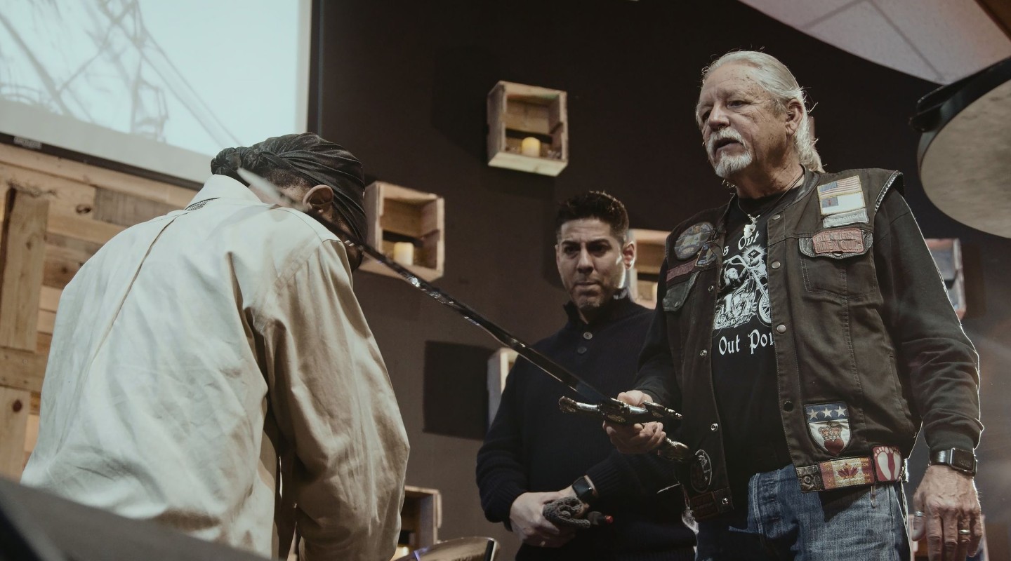Three individuals are standing in a triangle for what appears to be a knighting ceremony. One person is turned away from the camera, showing only their back. On top of their shoulder lays the end of a sword, held by the person across from them. That person is standing with a serious demeanor, dressed in a biker-style vest with various patches. The third person stands to the right of the biker, staring at the sword with a focused expression.