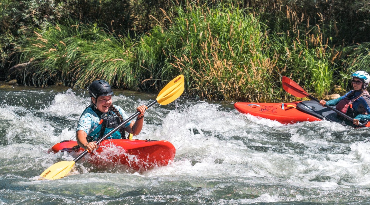 Two kayakers navigate rough waters, their expressions focused and exhilarated. The person in the foreground paddles vigorously, splashes surrounding their red kayak. The person behind, partially obscured by splashing water, also appears engaged in the challenge, maneuvering a red kayak with a blue top deck. The backdrop is full of lush greenery and vegetation.