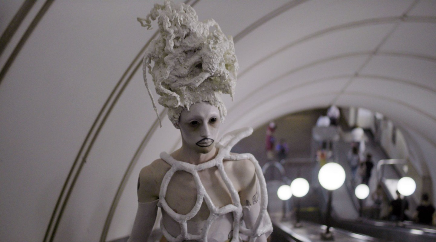 A figure with an avant-garde appearance is positioned in the foreground, dressed in what appears to be a white costume with structural, organic shapes enveloping the body and an elaborate headpiece resembling a stylized, textured wig or headdress. The figure's face is painted white, with darkened eyes and lips, and exhibits a neutral expression. The background shows an interior space with curved architecture and multiple people in motion, possibly a subway station or a public concourse, with a softly blurred focus that suggests movement and life happening around the still, striking figure in the foreground.
